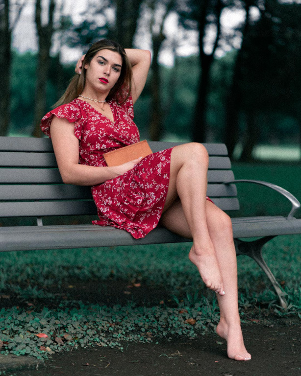 Fashion ediotrial photo of model posing on a park bench holding a book wearing a red dress